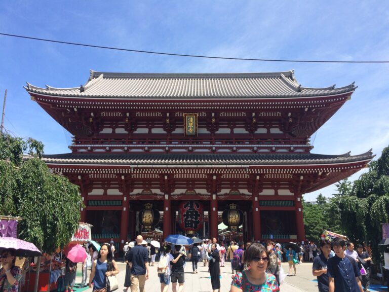 Visitors walk in front of the large, ornate red gate of Senso-ji Temple in Tokyo on a sunny day.