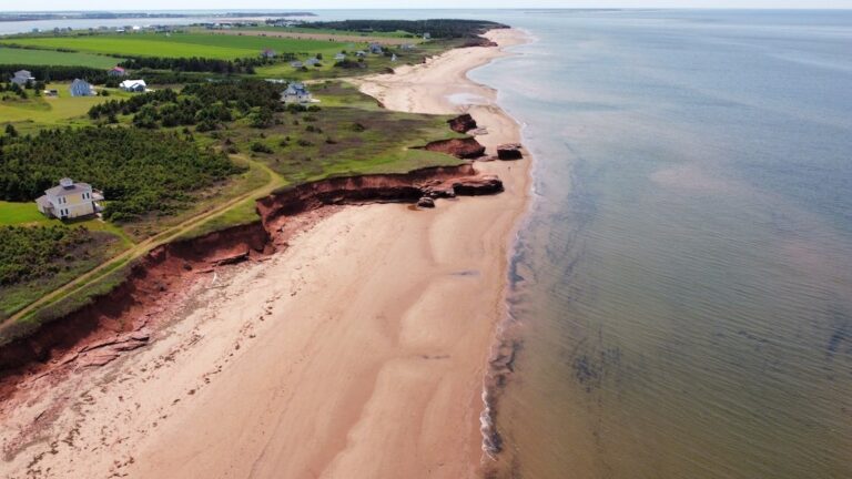 Aerial view of a coastal landscape with red cliffs, sandy beach, scattered houses, and calm ocean waters.