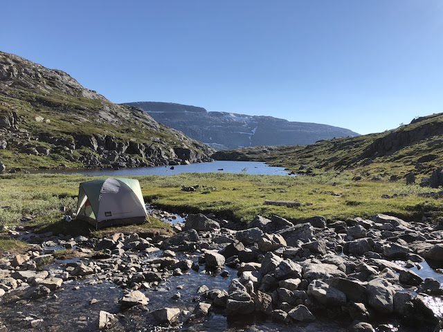 A tent is pitched on rocky ground near a small pond, surrounded by hills under a clear blue sky.