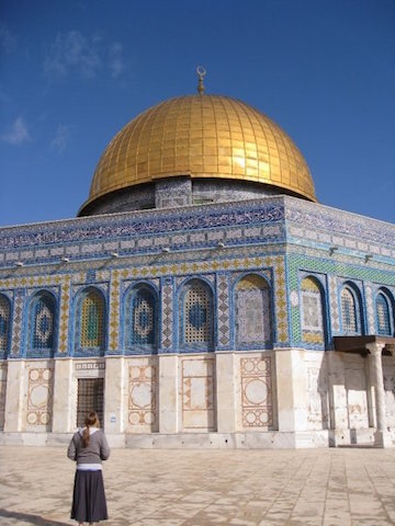 A person stands in front of the Dome of the Rock, featuring its iconic gold dome and intricate blue tilework under a clear sky.