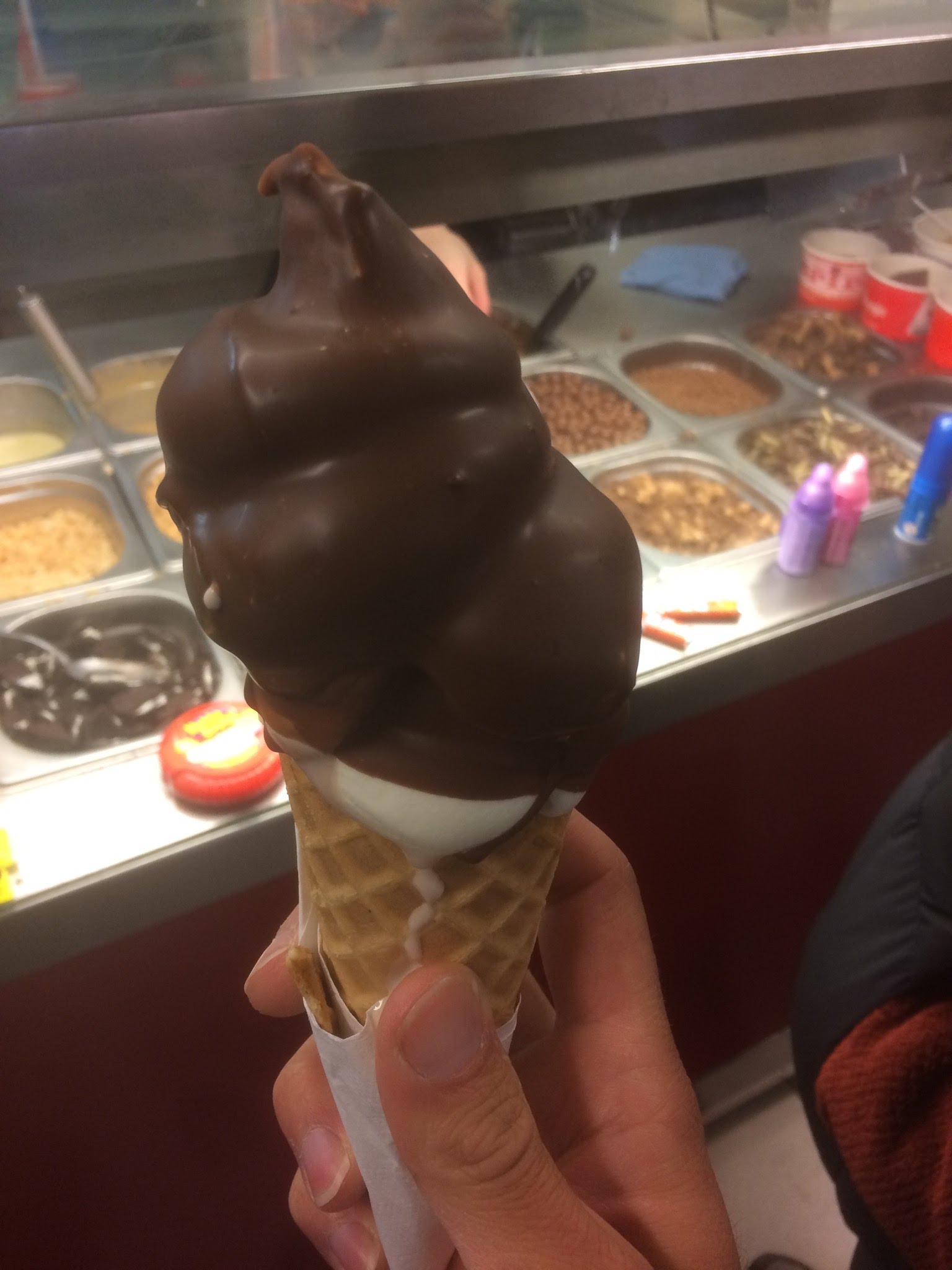 A hand holds a chocolate-dipped soft serve ice cream cone in front of a toppings bar at an ice cream shop.
