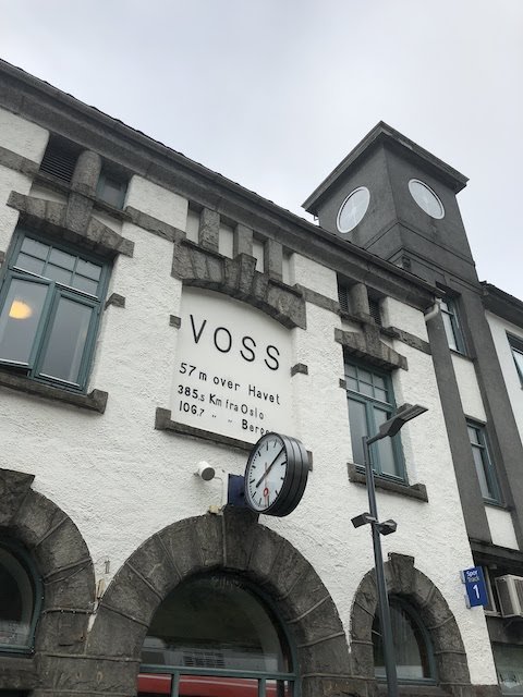 Exterior of Voss train station with a sign showing elevation and distances to Oslo and Bergen; clock and street sign visible.