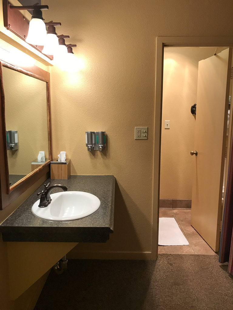 A bathroom with a wall-mounted sink, large mirror, soap dispensers, and an open door leading to a shower area with a white bath mat.