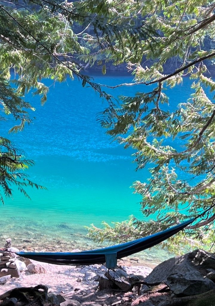 A hammock is tied between trees by the shore of a clear blue lake, with sunlight filtering through the branches.