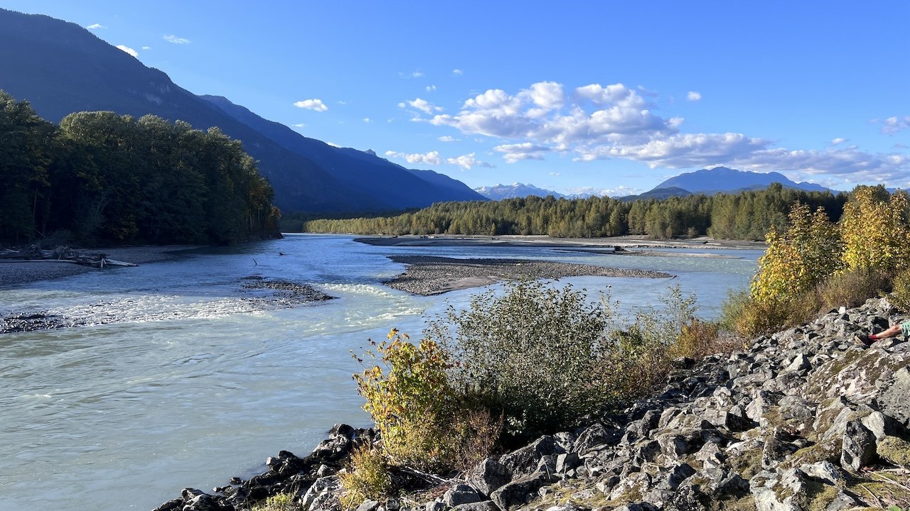Wide river flowing between forested banks and rocky shore with mountains in the background under a blue sky with scattered clouds.