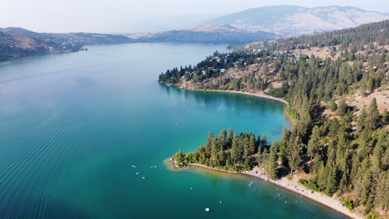 Aerial view of Okanagan lake with clear blue water, forested shoreline, and scattered houses on the hills in the background.