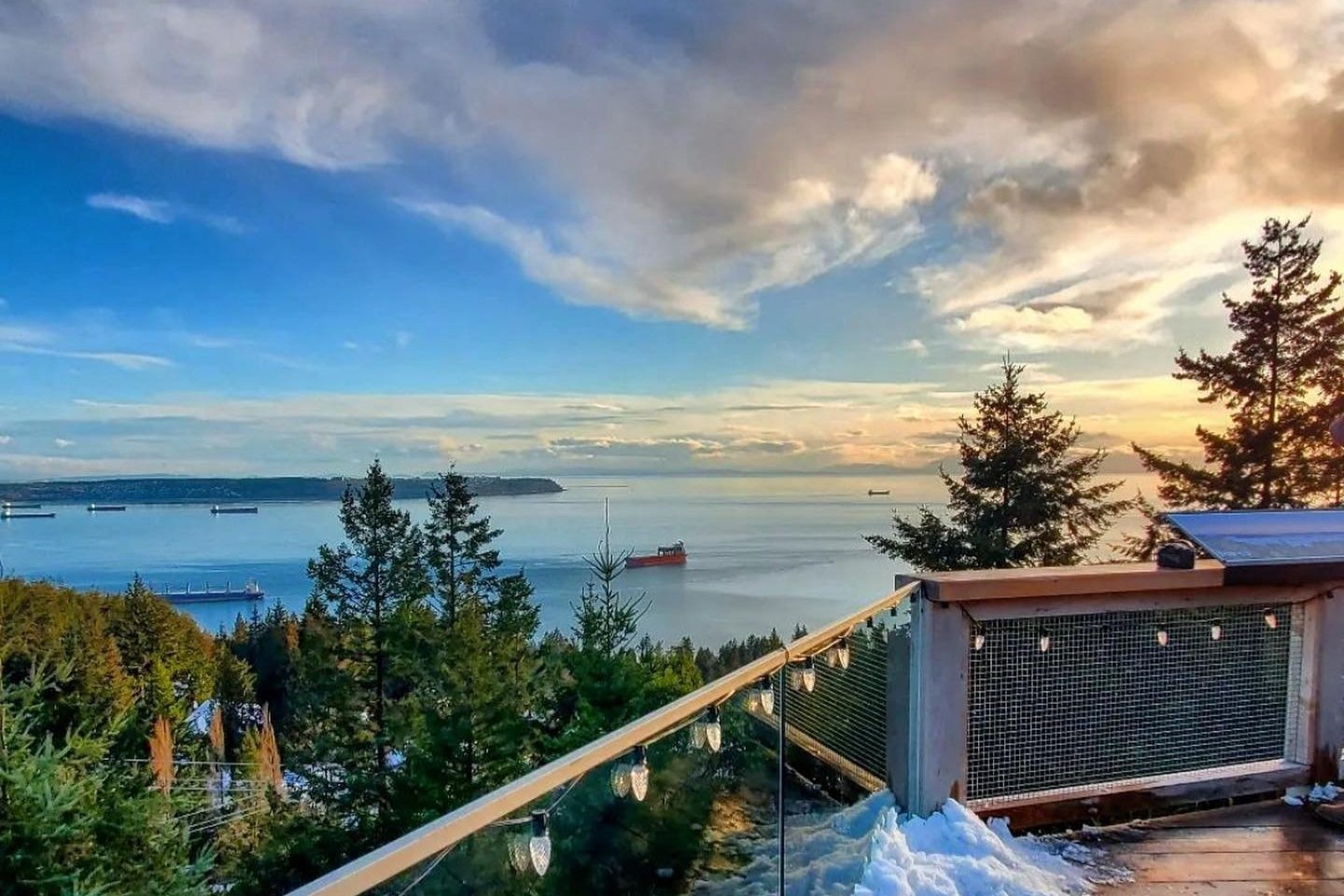 View of a calm sea with anchored ships, seen from a snowy deck surrounded by trees at sunset.