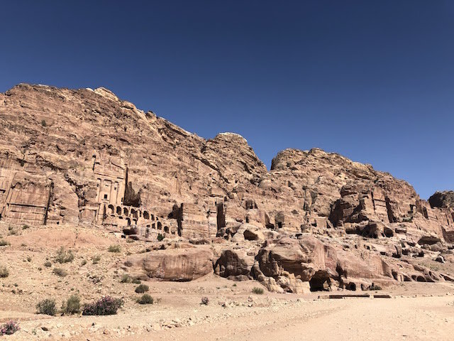 Ancient rock-cut structures and tombs are carved into a rugged sandstone cliff under a clear blue sky.