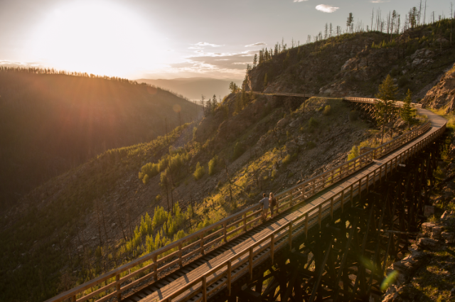A person walks on a wooden trestle bridge winding along a mountainside at sunset, with sunlight streaming over the hills.
