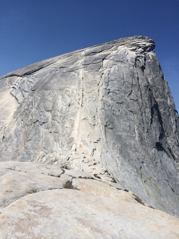 A steep granite rock face rises sharply under a clear blue sky, with a faint trail hinting at a hike up iconic Half Dome.