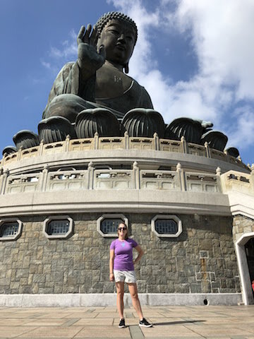 A person stands in front of a large seated Buddha statue on a stone platform, under a partly cloudy sky.