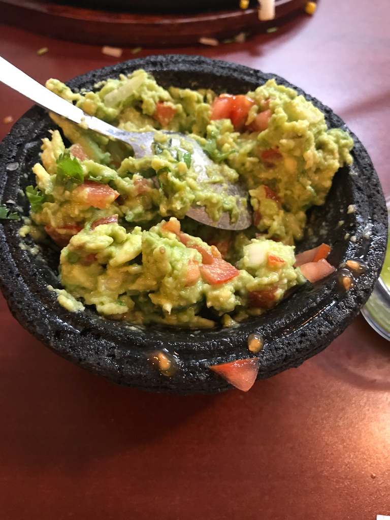 A stone bowl filled with chunky guacamole that contains tomatoes, onions, and herbs, with a spoon resting inside.