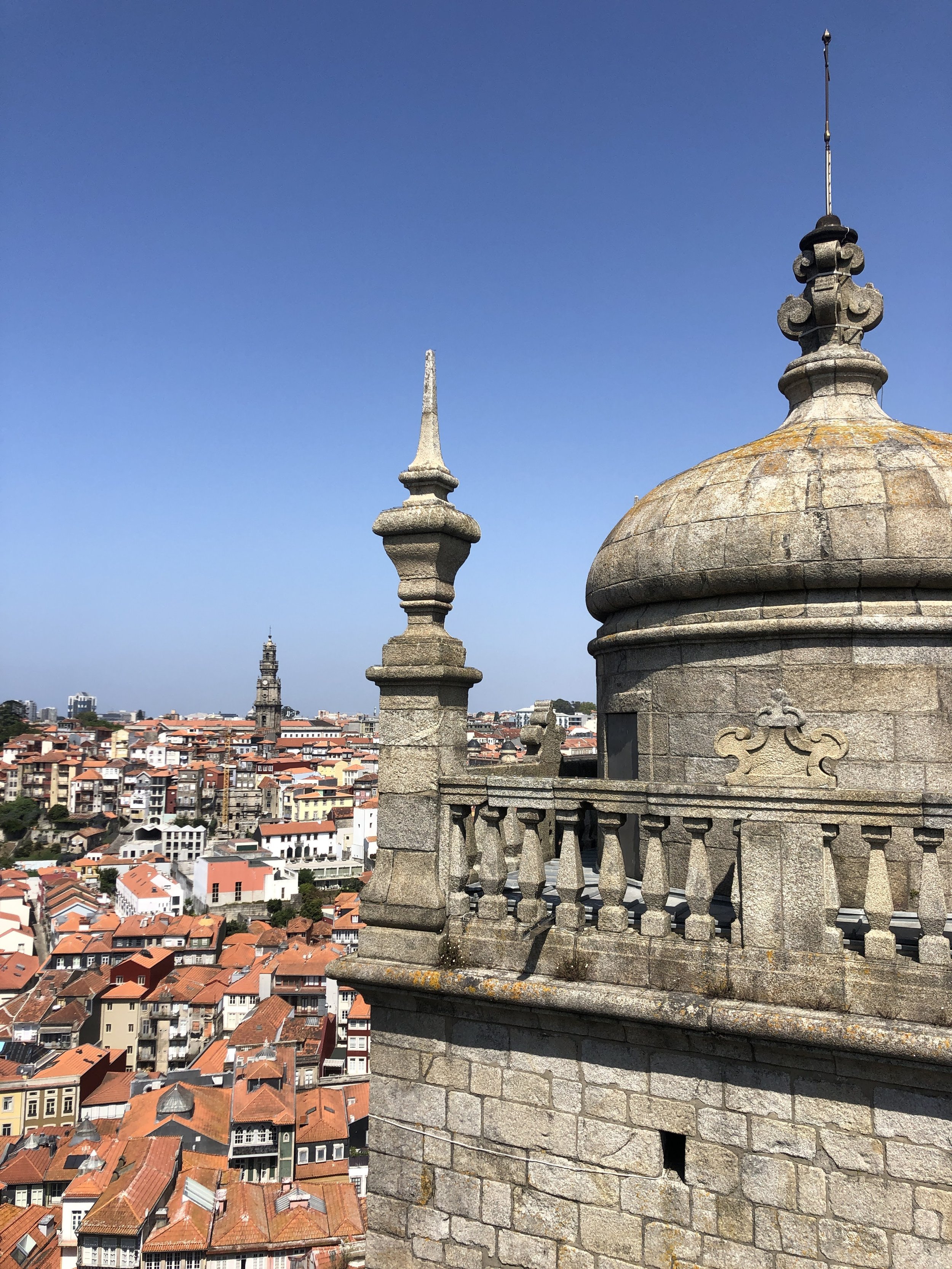 Stone turret with decorative spire overlooks a city with red-tiled rooftops under a clear blue sky.