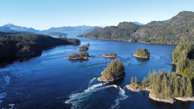 Aerial view of a coastal landscape with small forested islands, blue water, and surrounding tree-covered hills under a clear sky.