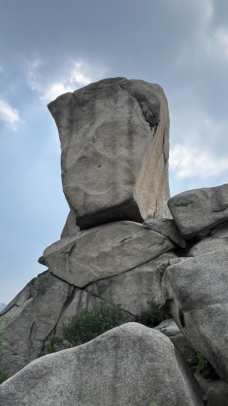 A large, upright boulder sits atop other rocks under a cloudy sky, with some greenery at the base.