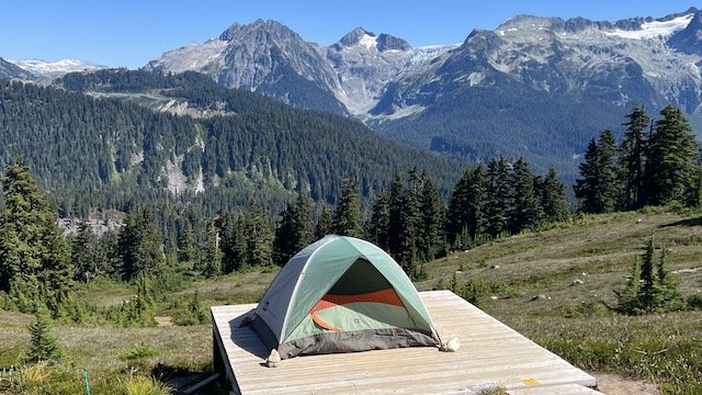 A green tent is pitched on a wooden platform in a mountain meadow, surrounded by pine trees and distant snow-capped peaks.