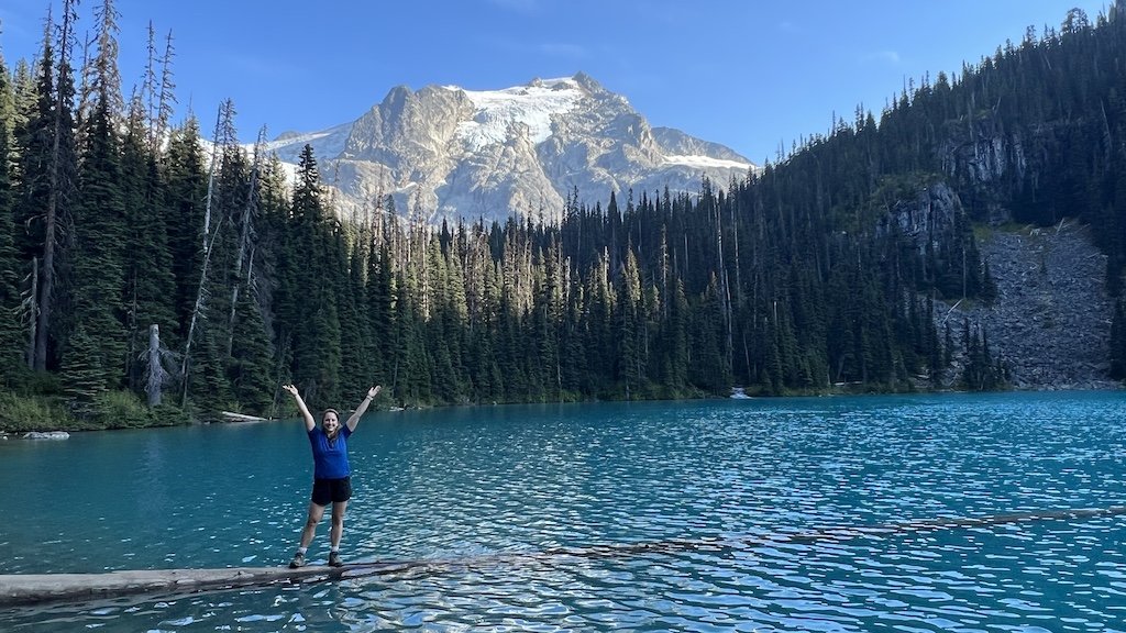 Person standing on a log in turquoise Joffre Lake, with pine trees and a snow-capped mountain, perfect for a scenic hiking adventure.