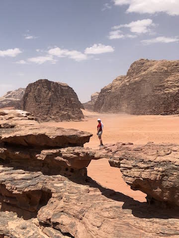 A person stands on a natural rock bridge in a desert landscape with rocky formations and a partly cloudy sky.