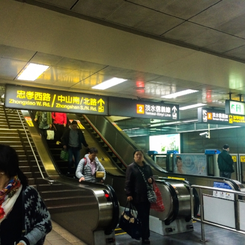 People use escalators in a subway station with bilingual signs directing to different lines and exits.