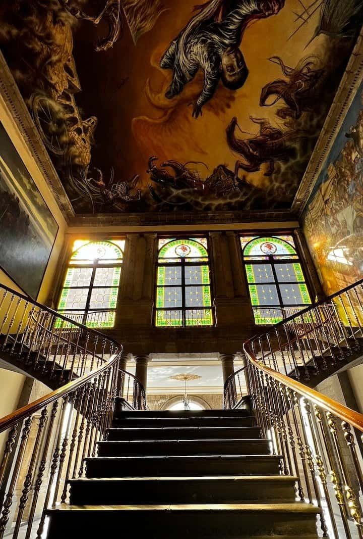 A grand staircase with ornate railings, stained glass windows, and a dramatic ceiling mural featuring a central human figure.