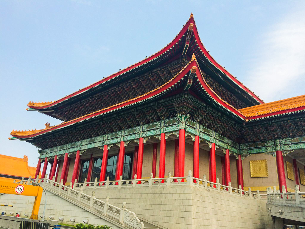 Large traditional Chinese-style building with ornate red columns, green and orange roof, and intricate decorative details under a clear sky.