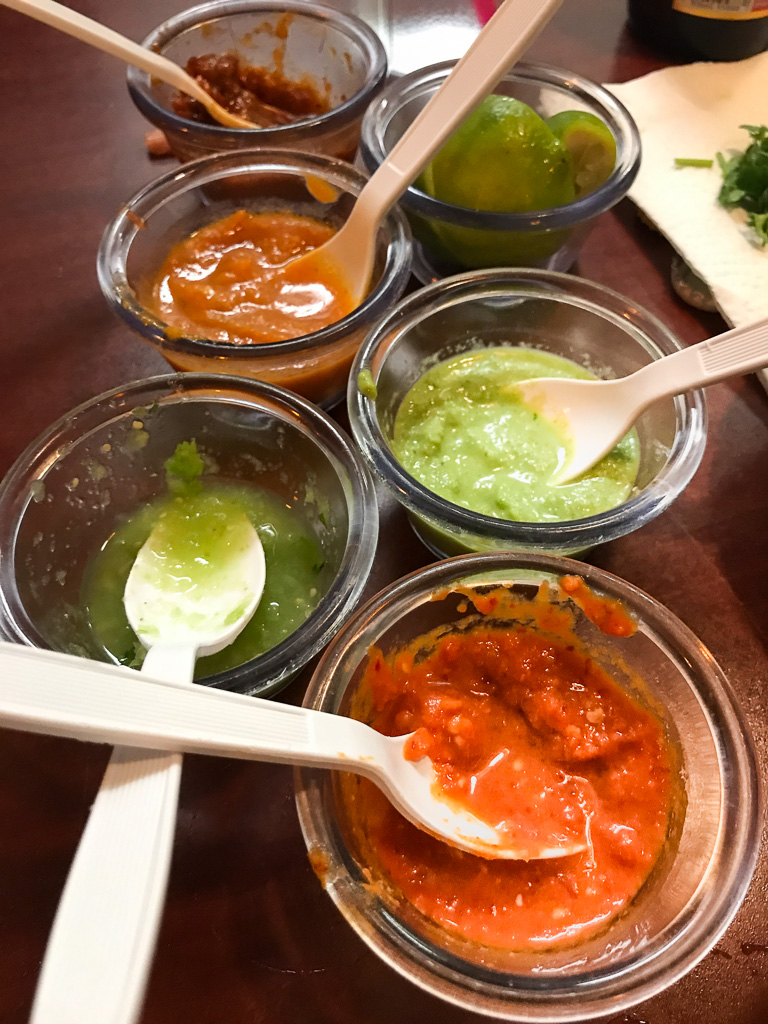 Six small bowls with different colored salsas and sauces, each with a white plastic spoon, are arranged on a wooden table.