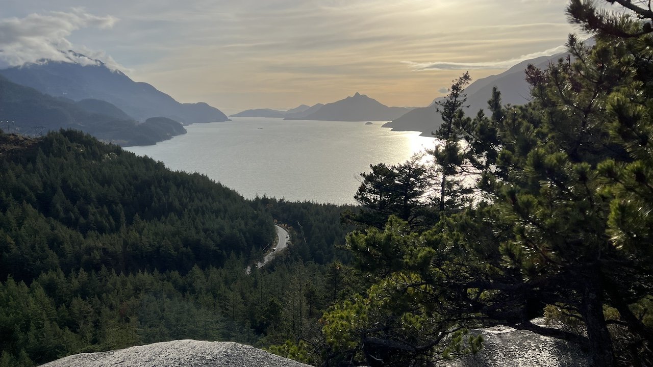 A winding road runs through a dense evergreen forest beside a large body of water, surrounded by mountains under a cloudy sky.