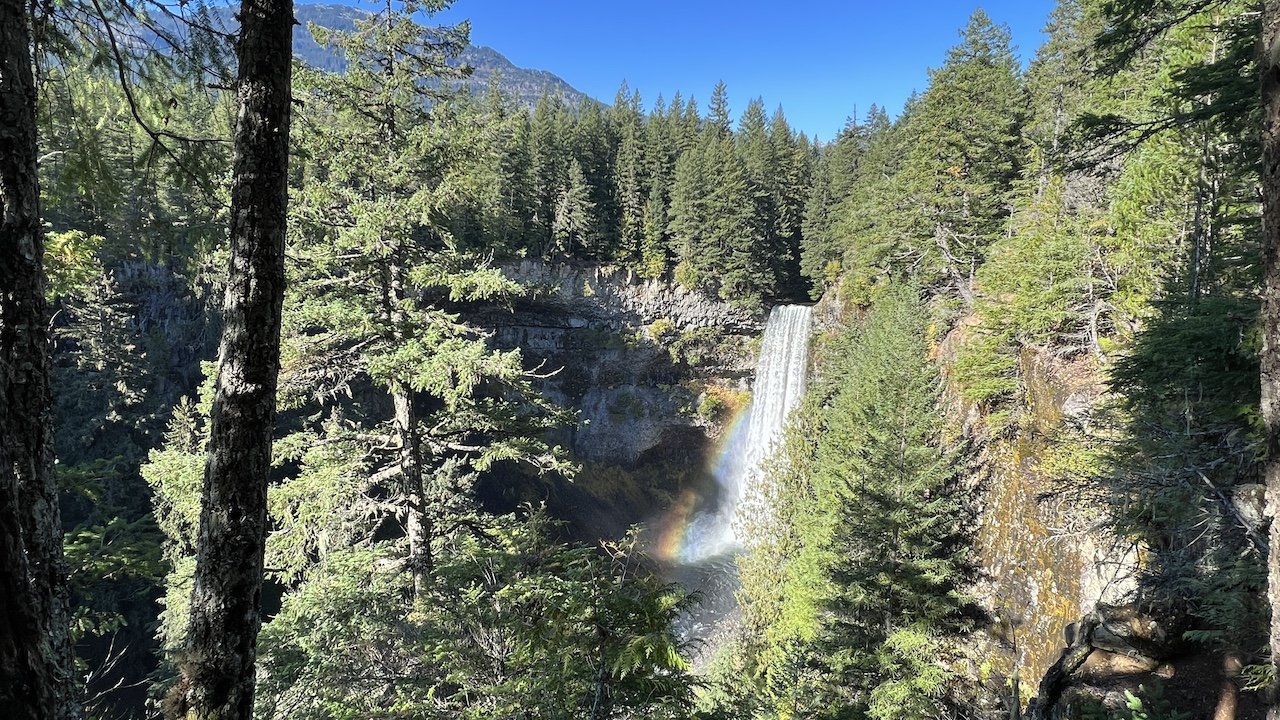 A forested landscape with a waterfall cascading into a ravine, a faint rainbow visible in the mist, under a clear blue sky.