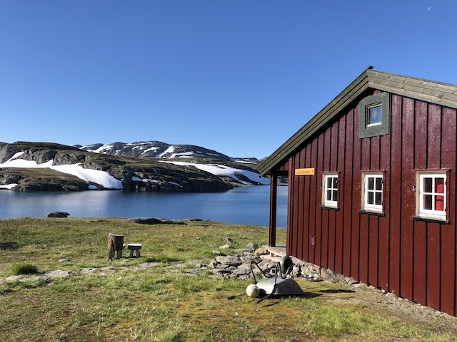 A red wooden cabin stands by a lake with snow-capped hills in the background under a clear blue sky.