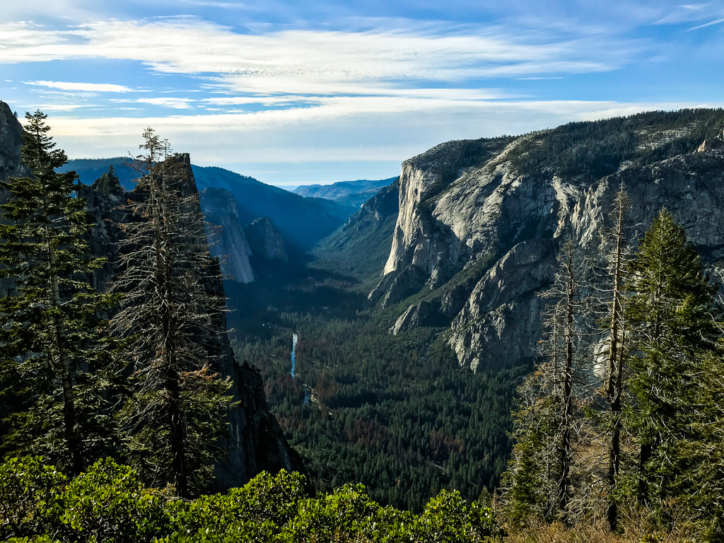 View of a forested valley surrounded by tall granite cliffs and distant mountains under a partly cloudy sky.