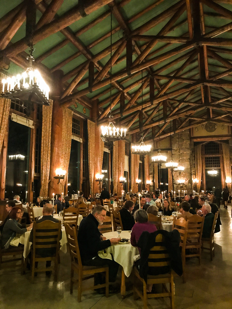 Large rustic dining hall with wooden beams, chandeliers, large windows, and people seated at tables eating dinner.
