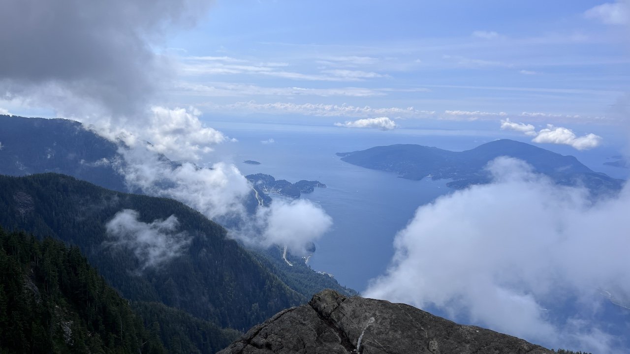View from a mountain summit overlooking a forested coastline, islands, and the ocean under a partly cloudy sky.
