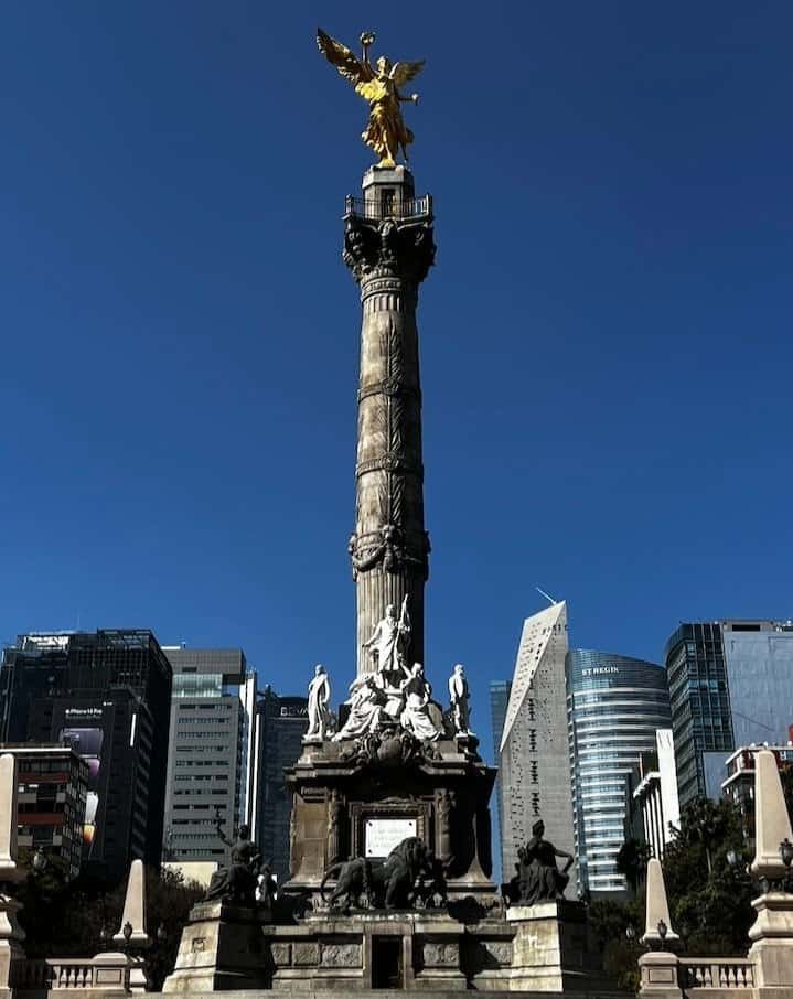 A tall stone column with a golden angel statue on top, surrounded by modern city buildings under a clear blue sky.