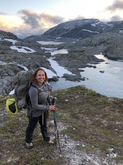 A woman with a large backpack and trekking poles stands on rocky terrain near a river and snow-capped mountains at dusk.
