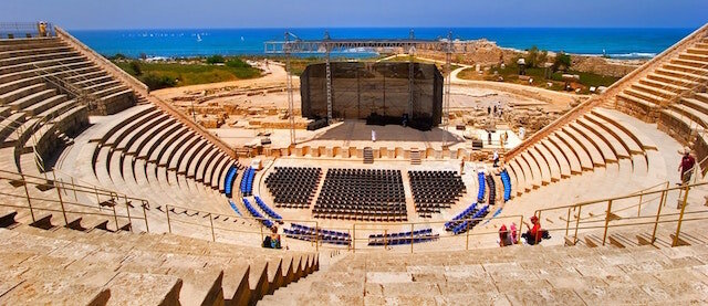 Ancient stone amphitheater with a central stage and chairs, located near the sea under a clear blue sky.