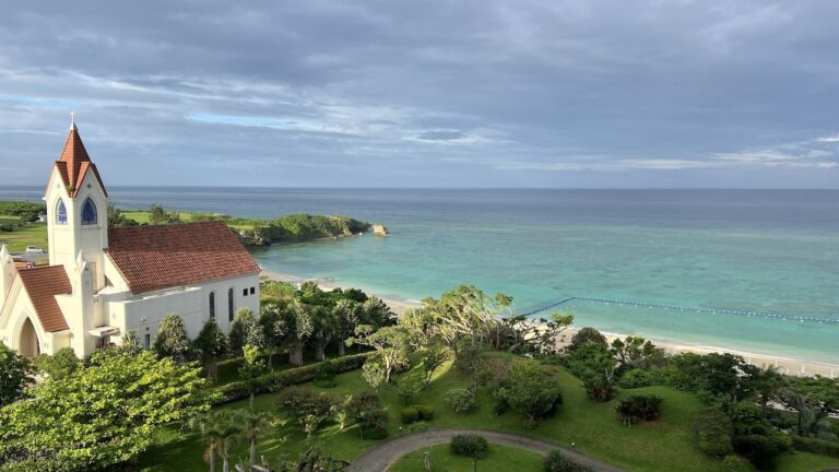 A white church with a red roof stands near a green garden overlooking a turquoise ocean under a cloudy sky.