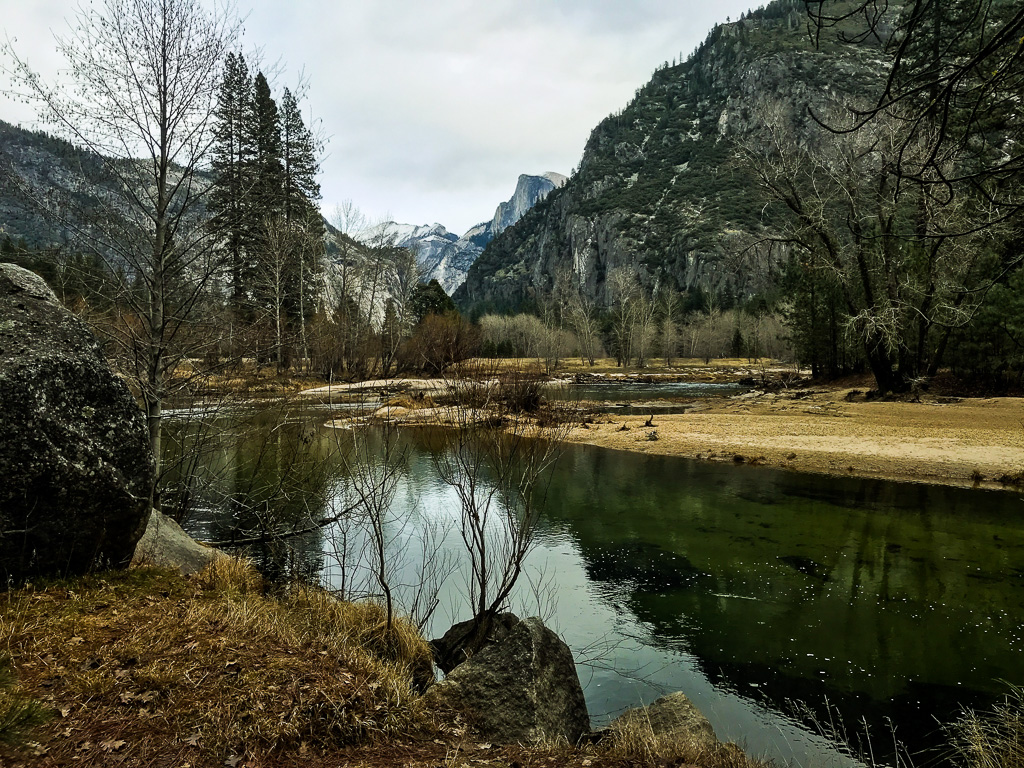 A calm river runs through a valley with rocky mountains and sparse trees under an overcast sky.