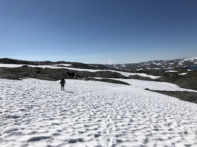 A person walks alone on a snow-covered landscape under a clear blue sky, with rocky hills and patches of snow in the background.