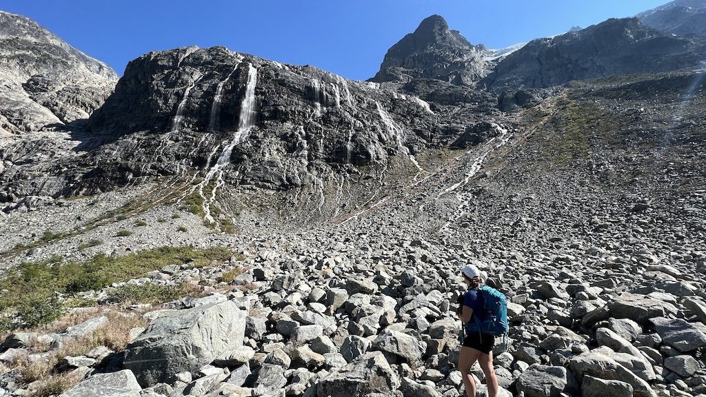 A person with a backpack stands on a rocky slope, facing a mountain with waterfalls under a clear blue sky.