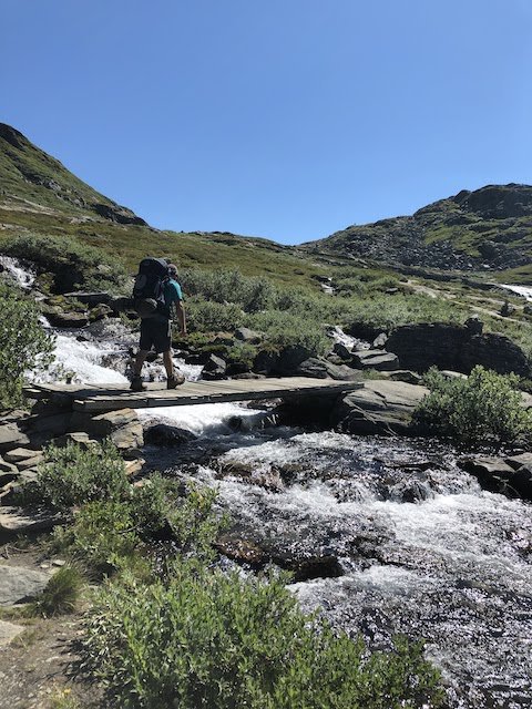 A hiker with a backpack crosses a small wooden bridge over a rocky stream in a mountainous landscape on a sunny day.