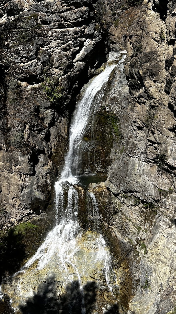 A waterfall flows down a rocky cliff surrounded by rugged stone surfaces and sparse greenery.