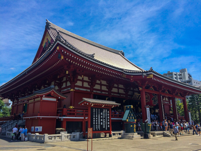 Large red wooden temple with ornate roof and columns, crowds of people at entrance, and blue sky overhead.