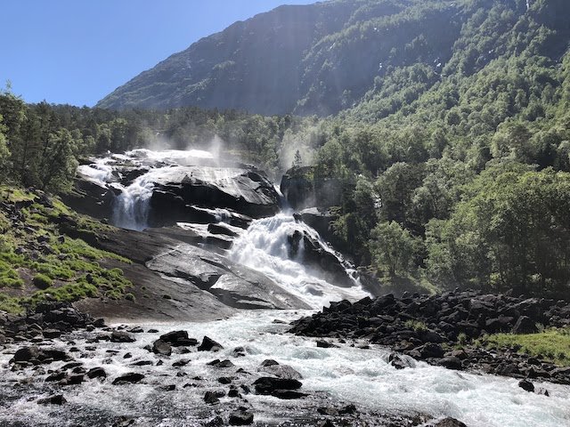 A waterfall cascades over dark rocks surrounded by green trees and mountains under a clear blue sky.