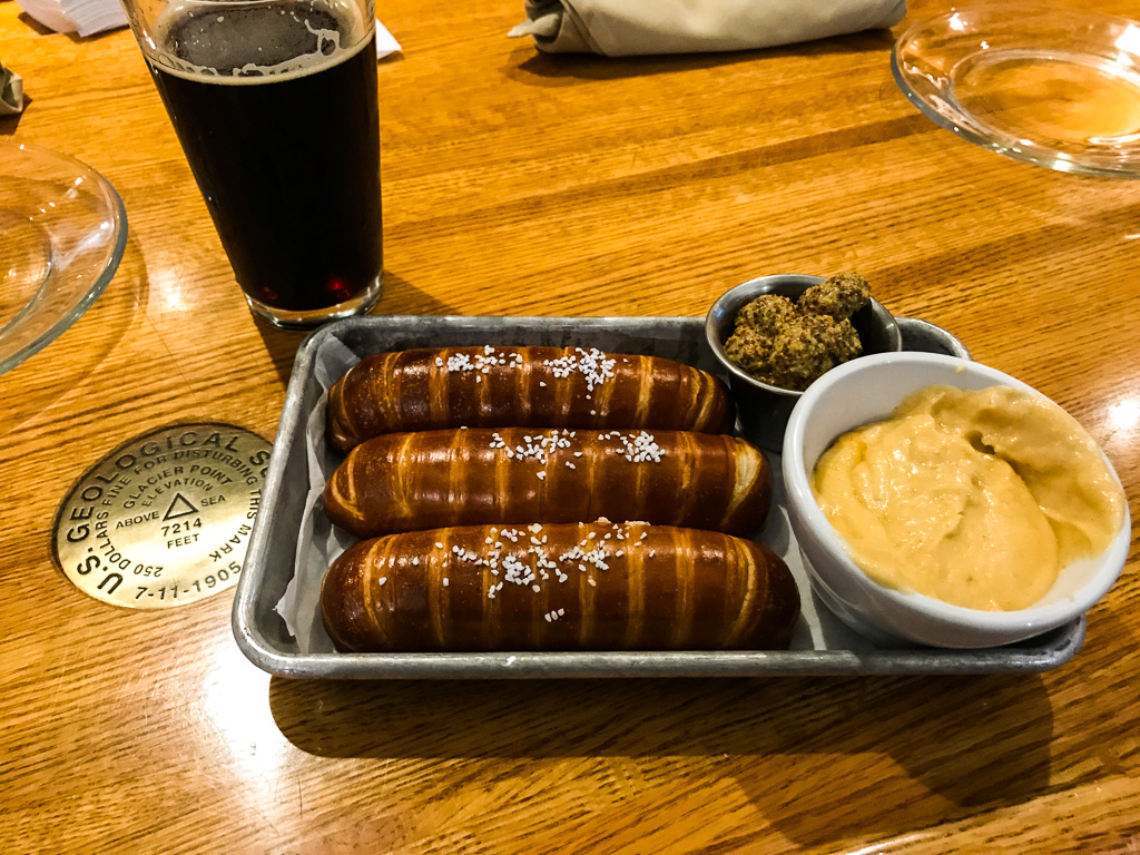 A metal tray with three pretzel sticks, a bowl of cheese dip, mustard, and a glass of dark beer on a wooden table.