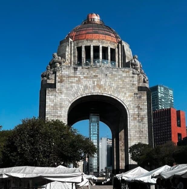 Large stone monument with an arched base and copper dome, surrounded by trees and tents under a clear blue sky.