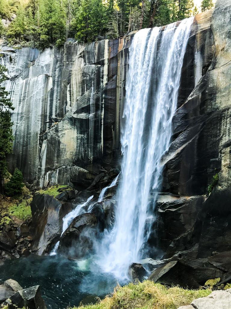 A tall waterfall cascades over a rocky cliff surrounded by trees, with water splashing into a pool below.