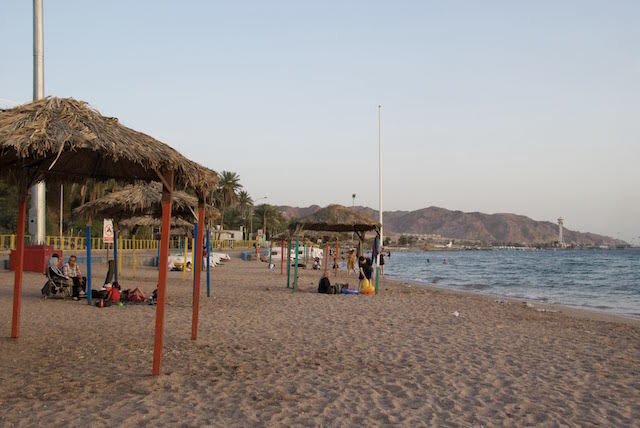 People relax under thatched umbrellas on a sandy beach near calm water, with distant hills and scattered palm trees in the background.