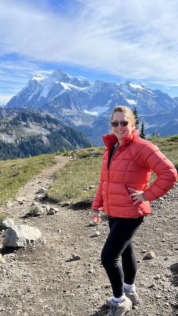 A woman in a red jacket stands on a mountain trail with snow-capped peaks and blue sky in the background.