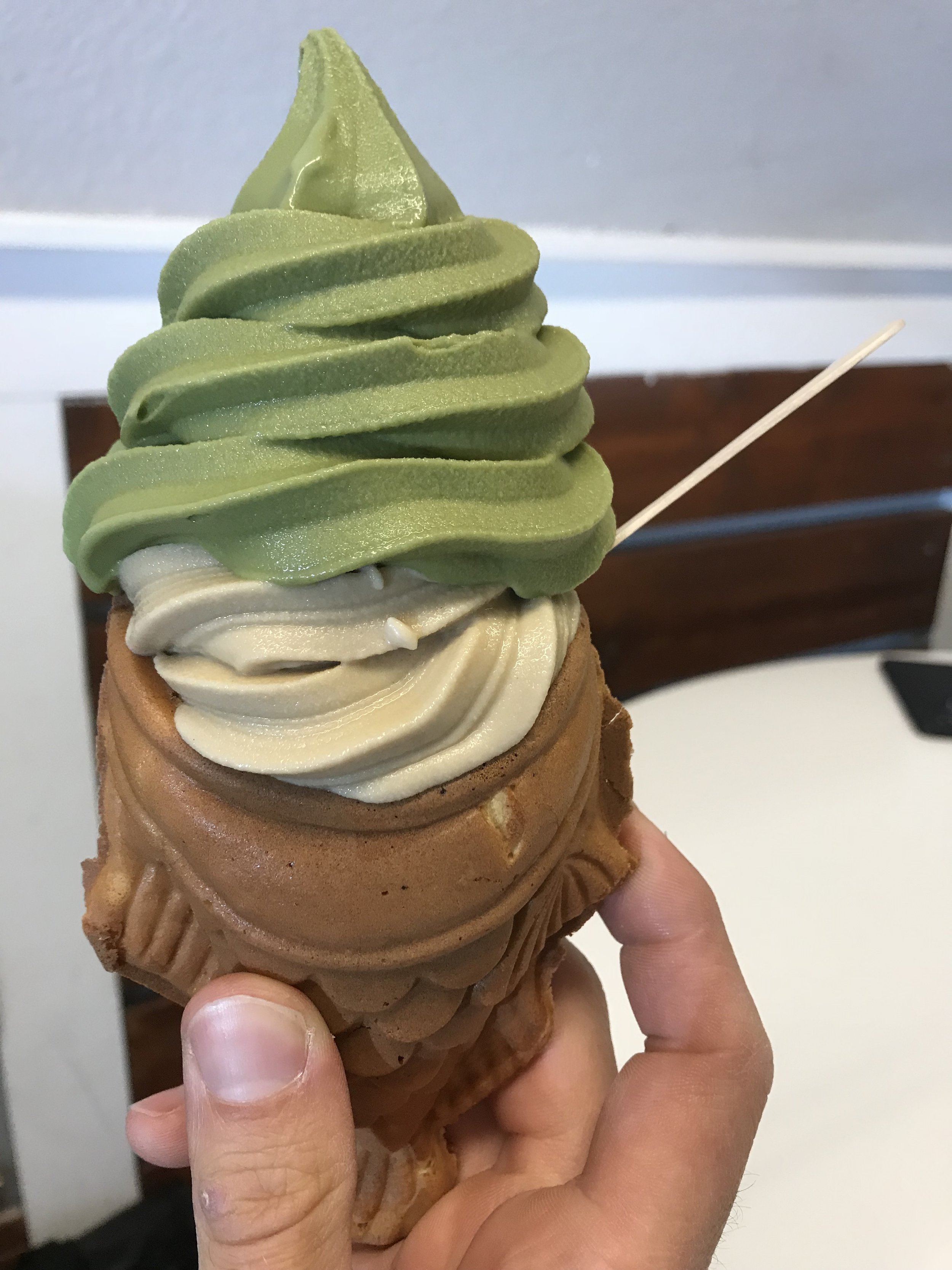 A hand holding a taiyaki ice cream cone with green tea and vanilla soft serve, and a wooden stick inserted on the side.