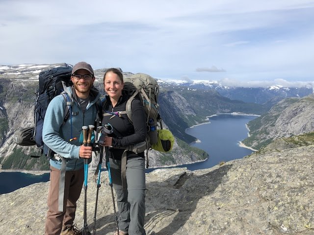 Two hikers with backpacks and trekking poles stand on a rocky cliff overlooking a fjord and mountains under a cloudy sky.